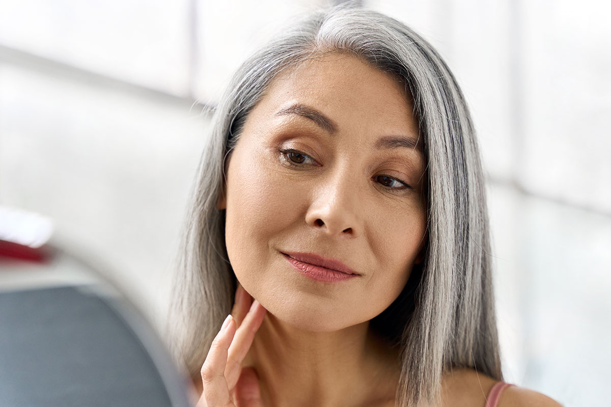 Asian lady with grey hair looking at her face in the mirror