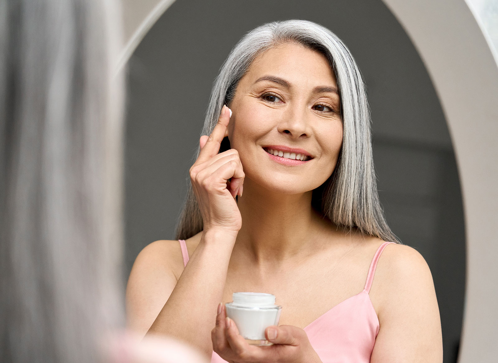 Older lady applying moisturizer on face in the mirror