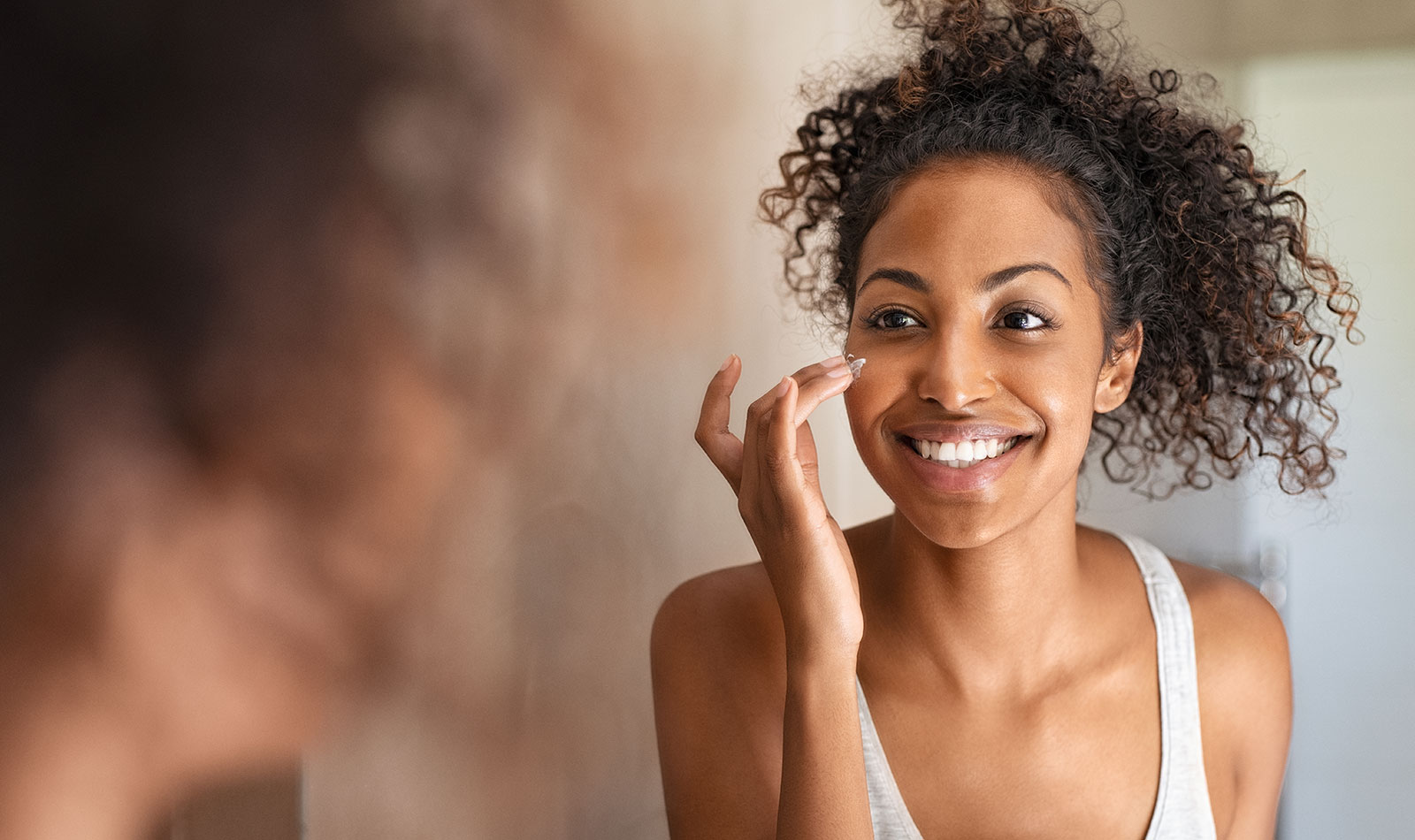 Young lady applying moisturizer on her face in the mirror